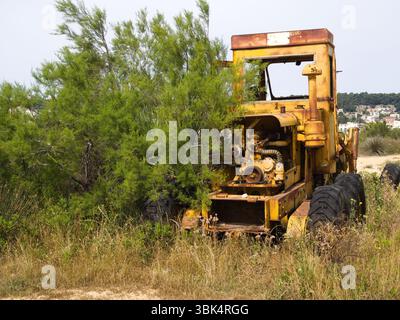 Tracteur jaune altéré avec des restes de moteur visibles abandonnés dans un champ de campagne sec à côté d'un buisson vert vibrant. Banque D'Images