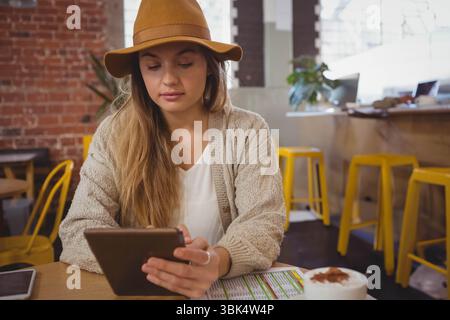 Femme assise dans un café en brique portant un chapeau à large bord tenant une tasse à café de smartphone de menu de tablette Banque D'Images