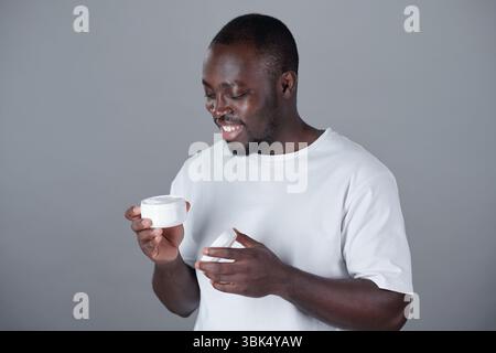 Homme afro-américain souriant tout en tenant un produit de soin de la peau sur fond gris. Vêtu d'une chemise blanche, il examine le produit et semble ravi Banque D'Images