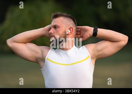 Un jeune homme en forme faisant des exercices d'étirement avant de courir ou de s'entraîner dans un parc, il écoute de la musique sur des écouteurs sans fil pour rester motivé. Actif Banque D'Images