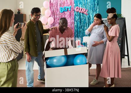 Homme noir émergeant d'une grande boîte remplie de ballons bleus tandis que la femme enceinte et les amis réagissent avec excitation Banque D'Images