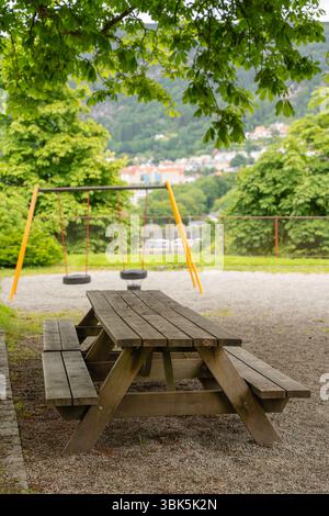 Table de pique-nique en bois vide près des balançoires de terrain de jeu dans le parc verdoyant à flanc de colline Banque D'Images