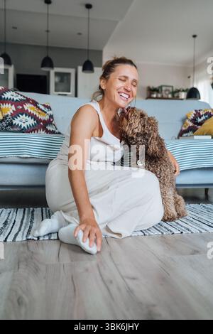 Femelle d'âge moyen riant joyeusement assis sur le sol stratifié du salon confortable embrassant le chien Maltipoo montrant la joie de se lier avec l'animal de compagnie bien-aimé. Banque D'Images