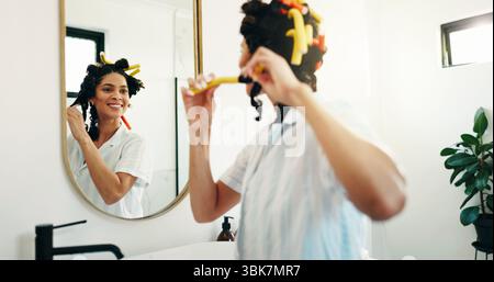Heureux, femme ou bigoudis dans le miroir pour se préparer, routine du matin ou coiffure pour le volume. Beauté, réflexion et fille à la maison avec soins capillaires Banque D'Images