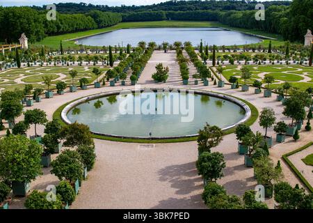 Vue aérienne du jardin formel, avec piscine circulaire, chemins de gravier, arbres en pot, pelouse bien entretenue, et lointain lac et forêt au Palais de Versaill Banque D'Images