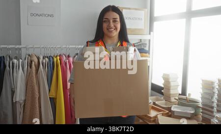 Une femme bénévole porte une boîte dans un centre de dons caritatifs entouré de vêtements et de contenants alimentaires à l'intérieur. Banque D'Images