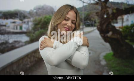 Blonde femme caucasienne enroule ses bras autour de ses épaules sur une promenade en bord de mer ; self-Care contentment relaxation paix. Banque D'Images