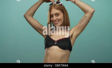 Jeune femme souriante avec des cheveux blonds portant des lunettes de soleil et un haut de bikini pose en toute confiance sur un fond vert vif isolé mettant en valeur le jeu Banque D'Images