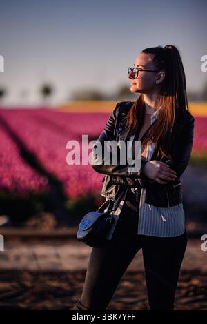 Femme élégante dans une veste en cuir et des lunettes pose devant des rangées vibrantes de tulipes sous la lumière dorée du soleil du soir. Banque D'Images