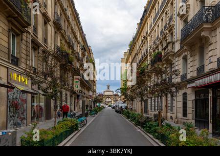 Vue sur la rue avec des bâtiments bordant la route menant à une arche au loin, avec un ciel nuageux. Banque D'Images