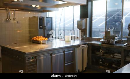 Cuisine moderne du restaurant avec comptoir en acier inoxydable, oranges fraîches dans un plateau, grandes fenêtres et éclairage bokeh flou de la hauteur Banque D'Images