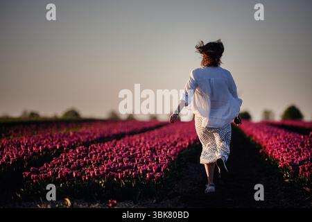 Vue arrière d'une femme marchant entre des rangées de tulipes roses pendant l'heure dorée, entourée par la nature et la lumière chaude. Banque D'Images