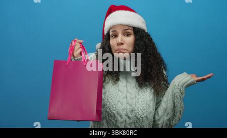 Femme portant le chapeau de père noël et le pull tient le sac à provisions rose avec l'expression confuse devant le fond bleu indiquant le dilemme du shopping de vacances. Banque D'Images