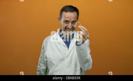 Homme en manteau blanc tenant un aligneur dentaire sur fond orange, exsudant la confiance et l'expertise, professionnel mature présentant l'orthodo de pointe Banque D'Images
