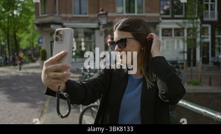 Femme prenant selfie et souriant avec smartphone à amsterdam, marchant le long de canaux pittoresques dans une tenue élégante pendant une journée ensoleillée mettant en vedette l'emblématique c hollandais Banque D'Images
