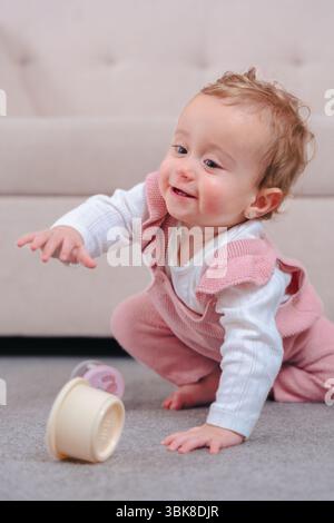 Happy Baby girl en salopette rose s'agenouille sur le tapis, atteignant pour attraper le jouet roulant Banque D'Images
