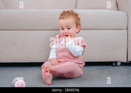 Bébé fille en salopette rose assise sur la moquette, dentition sur ses doigts Banque D'Images