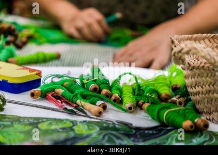 Les bobines de trame (en français flûtes) en laine vierge verte sont posées sur un tissu blanc à côté d'une tapisserie de grande taille inspirée d'un motif de l'artiste japonais Hayao Miyazaki tiré de "Mon voisin Totoro" dans l'atelier créatif de la Cité internationale de la tapisserie sur la Rue des Arts à Aubusson, Creuse, Nouvelle-Aquitaine, France. Le motif scintille à travers les fils de trame. Derrière, les mains de la tisserande Aurélie travaillent sur la trame. Une paire de ciseaux à manche rouge, un long outil métallique et une boîte jaune et bleue sont posés à côté des bobines, et un panier tissé se trouve à droite. Une impression du motif en nuances o Banque D'Images