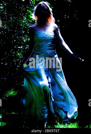 Une femme dans une robe bleue chatoyante se tient dans une forêt sombre, les bras tendus. Banque D'Images