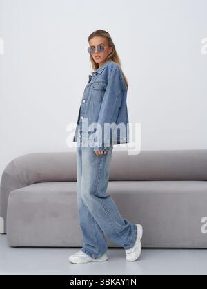 Une jeune femme se tient confiante dans une veste en denim oversize élégante et un Jean ample, portant des lunettes de soleil réfléchissantes et des baskets blanches, posées à côté Banque D'Images