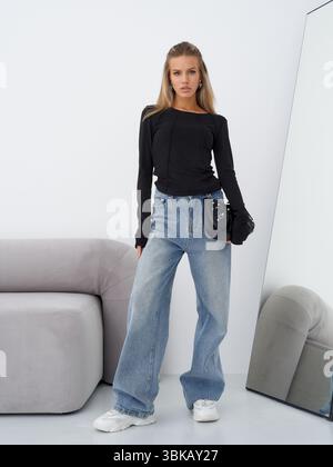 Femme décontractée avec de longs cheveux se tient en toute confiance à côté d'un canapé, tenant un petit sac. Elle porte un haut noir, un Jean large bleu et du blanc Banque D'Images