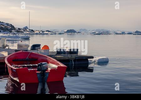 Un bateau à moteur rouge amarré parmi la glace de mer à Saqqaq, Groenland. Les collines enneigées et les fjords reflètent les conditions printanières arctiques et la vie active rurale. Banque D'Images