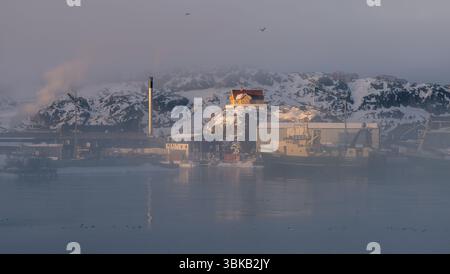 Port industriel et maison colorée au sommet d'une colline à Sisimiut, au Groenland, avec la brume arctique, la lumière dorée et la fumée s'élevant des infrastructures urbaines. Banque D'Images