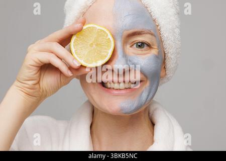 Femme heureuse appliquant un masque facial en argile et tenant du citron, appréciant raffermissant, hydratant et anti-âge soins de la peau à la maison. Banque D'Images