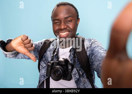 POV de photographe masculin debout et ludique donnant un pouce vers le bas sur fond isolé. Vlogger afro-américain avec appareil photo reflex numérique et sac à dos s'enregistrant lui-même alors qu'il fait des gestes de désaccord. Banque D'Images