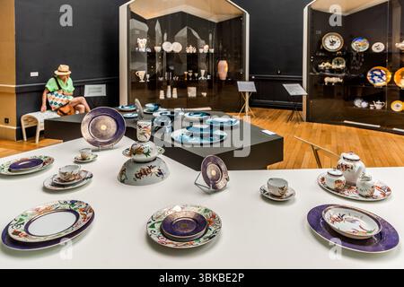 Une exposition au Musée National Adrien Dubouché sur la Place Winston Churchill à Limoges, Haute-Vienne, Nouvelle-Aquitaine, en France, présente des séries de porcelaine de marques bien connues telles que Bernardaud (anciennement Manufacture Royale de Limoges), Haviland, Jacques Pergay, Raynaud et Ateliers Arquié. Sur une table blanche, des couverts sont disposés : des assiettes avec des bords violets et dorés, des assiettes et des tasses avec des motifs floraux colorés, ainsi qu’une théière avec une cruche à lait et un sucrier. Derrière eux, des assiettes bleues reposent sur une plateforme sombre, et les vitrines contiennent des assiettes murales, des figurines et des vases. La journaliste Angela Berg est assise sur un Banque D'Images