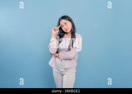 Une femme se tient debout devant un mur bleu, portant une veste rose et un pantalon blanc. Elle regarde en bas et elle est profondément dans la pensée Banque D'Images