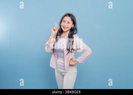 Une femme est debout devant un mur bleu avec une chemise rose et un pantalon blanc. Elle pointe son doigt vers le haut et sourit Banque D'Images