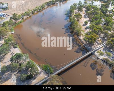 Vue aérienne d'un pont de suspension pour piétons au-dessus d'un coude de rivière brun à Horsham dans le Victoria, Australie. Banque D'Images