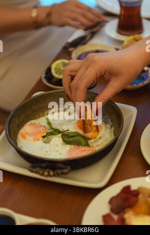 Un gros plan de la main d'une femme trempant du pain dans des œufs frits sur une table en bois pour le petit déjeuner dans un hôtel. Moment du matin confortable et appétissant. Banque D'Images