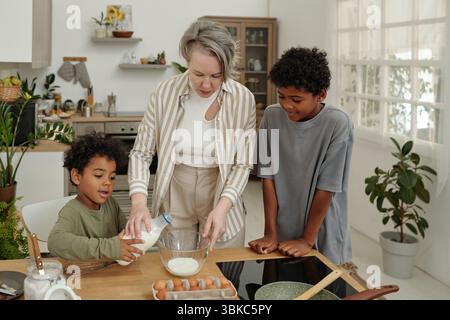 Femme guidant deux enfants dans l'activité de cuisson à l'intérieur d'une cuisine élégante avec des éléments en bois et métalliques et une lumière naturelle brillante. Cuisine remplie de divers ingrédients de cuisson autour Banque D'Images