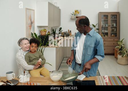 Famille multiethnique appréciant la cuisson des crêpes dans la cuisine de la maison. Femme d'âge moyen tenant l'enfant tandis que l'homme en chemise en denim préparant des ingrédients sur le comptoir de cuisine Banque D'Images