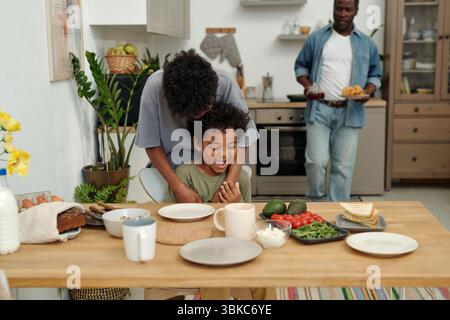 Réunion de famille dans la cuisine moderne avec la mère embrassant l'enfant et le père tenant la plaque de nourriture. Divers ingrédients frais et plats affichés sur la table en bois Banque D'Images