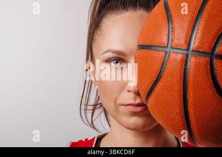 Gros plan portrait d'une joueuse de basket-ball déterminée, à moitié le visage couvert par le ballon, symbolisant la concentration et l'ambition. Banque D'Images