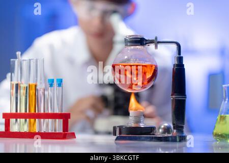 Une fiole de liquide rouge est chauffée au-dessus d'une flamme sur une paillasse de laboratoire remplie d'équipement. En arrière-plan, un scientifique flou travaille, créant une scène de A. Banque D'Images