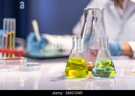 Gros plan de liquides chimiques colorés dans des fioles Erlenmeyer sur une table de laboratoire, avec un scientifique travaillant en arrière-plan, mettant l'accent sur les R scientifiques Banque D'Images