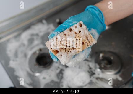 Vue de dessus du travailleur de l'entreprise de nettoyage dans des gants pressant l'eau savonneuse de l'éponge pendant le nettoyage du poêle à gaz sale, pour enlever la saleté tenace et la graisse Banque D'Images