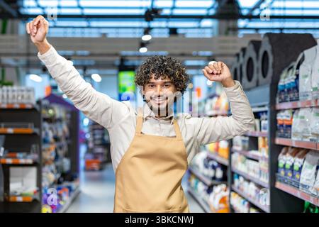 Un employé de magasin joyeux avec les cheveux bouclés fait la fête avec les bras levés, souriant largement dans une allée de supermarché. Banque D'Images