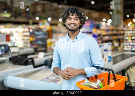 Un homme souriant avec les cheveux bouclés dans une épicerie, tenant un panier. Banque D'Images