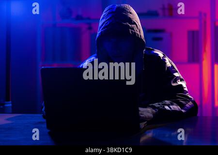 Un jeune homme portant un sweat à capuche noir utilisant intentionnellement un ordinateur portable dans une pièce éclairée au néon, évoquant les thèmes de la cybersécurité, du piratage, du codage et de la confidentialité numérique dans un t Banque D'Images