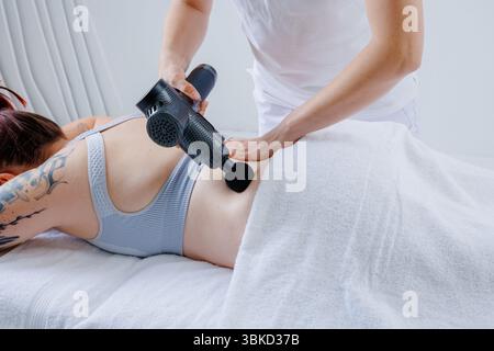 Massothérapeute appliquant un pistolet à percussion aux clients dans le bas du dos pour soulager la douleur musculaire, réduire la tension et soutenir la récupération du bien-être spinal. Banque D'Images