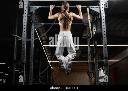 Vue arrière d'une femme en forme faisant des pull-ups sur un bar dans une salle de gym. Des muscles définis et une posture athlétique soulignent la force et la discipline dans l'entraînement physique. Banque D'Images