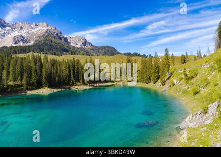 Les eaux turquoises vibrantes du lac Carezza entourées de forêts de pins verdoyantes et de collines ondulantes, avec les Dolomites enneigées en arrière-plan Banque D'Images