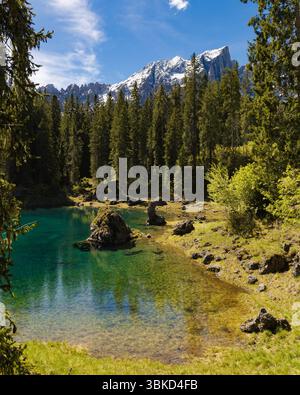 Les eaux turquoises claires et vibrantes du lac Carezza dans les Dolomites, entourées d'une dense forêt de pins et d'un rivage rocheux, avec des montagnes enneigées Banque D'Images