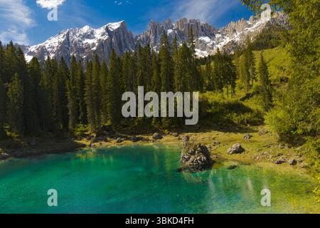Les eaux turquoises vives du lac Carezza dans les Dolomites, encadrées par une dense forêt de pins et les sommets enneigés et déchiquetés des montagnes sous A. Banque D'Images