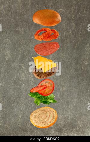 Hamburger volant avec ses ingrédients laitue pain tomates salami au fromage de bœuf et oignons croustillants sur fond gris. Banque D'Images